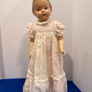 May include: A vintage doll with a painted face, wearing a light pink dress with eyelet details and ruffled trim. The doll has short, styled hair and is posed standing upright. The dress features a delicate, openwork design.