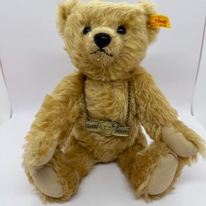 Peut inclure: Un ours en peluche brun clair avec des yeux et un nez noirs. Il porte une étiquette métallique avec les années 1902 et 2002. Une étiquette jaune est attachée à son oreille.