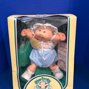 Op de afbeelding: Een Cabbage Patch Kids Preemie pop in de originele gele kartonnen verpakking. De pop heeft een zacht perzikkleurig lichaam, draagt een wit mutsje met kant, een topje met patroon, een blauwe pofbroek en witte schoentjes.