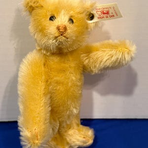 May include: A golden-yellow teddy bear with a tag that reads "Made in Germany by Steiff". The plush toy has a classic design with a stitched nose, black eyes, and jointed limbs. The bear is standing on its hind legs with one arm extended.