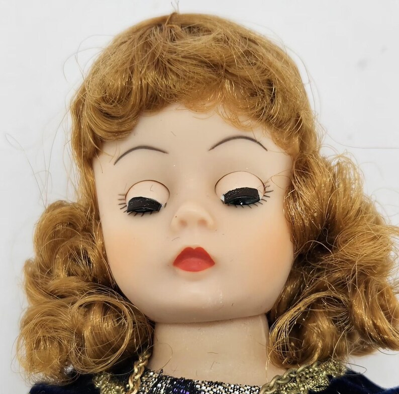 May include: A vintage doll with closed eyes, red lips, and auburn hair styled in curls. The doll has a porcelain face with painted features and is wearing a dark blue dress with gold trim. A gold necklace is visible.