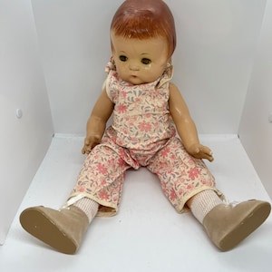 Könnte beinhalten: Eine antike Porzellanpuppe mit braunen Haaren, die ein Kleid mit Blumenmuster und braune Schuhe trägt. Die Puppe sitzt auf einer weißen Oberfläche.