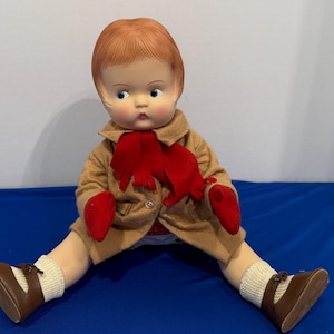 May include: A vintage doll with red hair, blue eyes, and a neutral expression. The doll wears a tan coat, red scarf and mittens, white socks, and brown shoes. The doll is sitting with its legs spread.