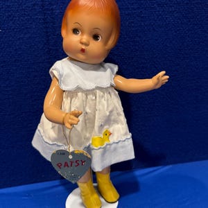 Könnte beinhalten: Eine Vintage-Puppe mit roten Haaren, die ein weißes Kleid mit blauen Verzierungen und gelbe Stiefel trägt. Die Puppe hält ein herzförmiges Schild mit der Aufschrift "Ich bin Patsy".