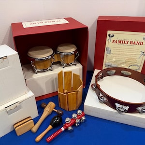 May include: A Family Band musical instrument set including a pair of wooden bongo drums, a brown tambourine with metal jingles, a wooden block, a wooden mallet, a black maraca, and a red handle with metal bells, all packaged in red and white boxes.