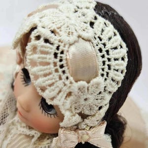 May include: A vintage doll with dark hair and large eyes, wearing a cream-colored lace bonnet with a matching bow. The bonnet is intricately crocheted, adding a delicate touch to the doll's appearance. The doll's face has a serene expression.