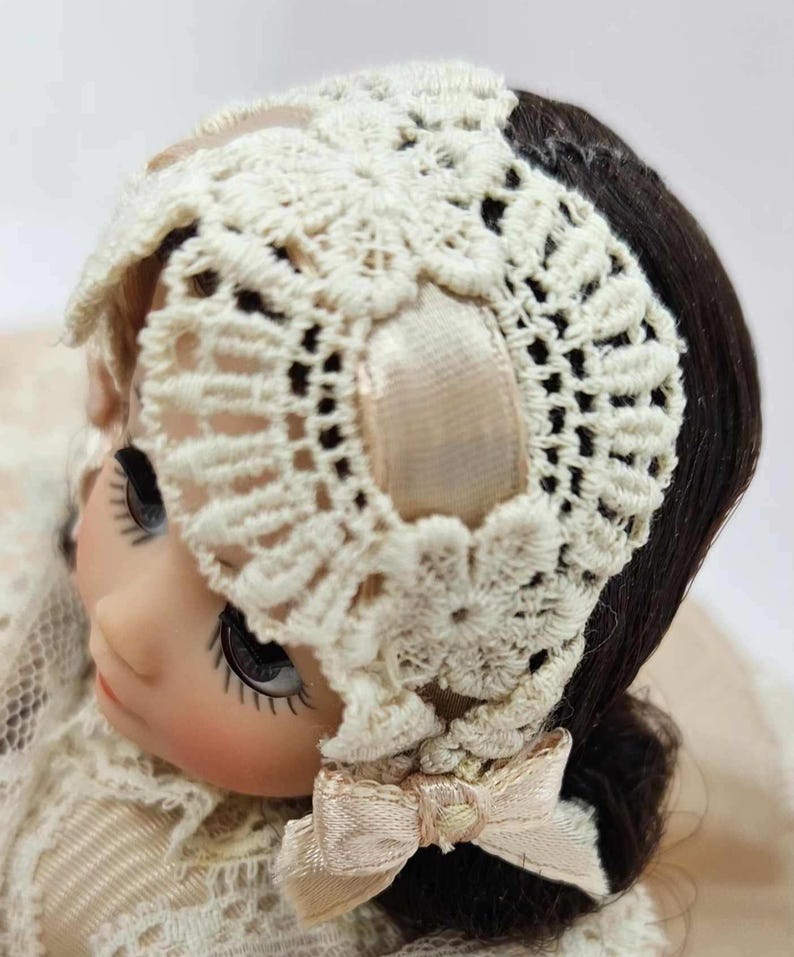 May include: A vintage doll with dark hair and large eyes, wearing a cream-colored lace bonnet with a matching bow. The bonnet is intricately crocheted, adding a delicate touch to the doll's appearance. The doll's face has a serene expression.