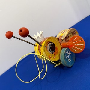 May include: A vintage wooden pull-along toy bee. The bee is painted yellow with black stripes, a smiling face, and a small white crown. It has red antennae and a yellow pull string. The wings are orange and yellow. The wheels are blue and orange.