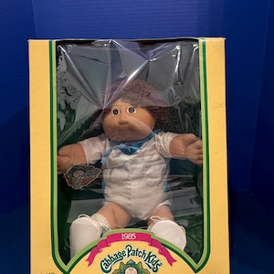 Könnte beinhalten: Eine 1985 Cabbage Patch Kids Puppe mit braunen Haaren und braunen Augen, bekleidet mit einem weißen Outfit mit blauem Kragen. Die Puppe befindet sich in einem gelb-grünen Karton mit dem Text Cabbage Patch Kids, 1985, und I'm unique. I'm adoptable.