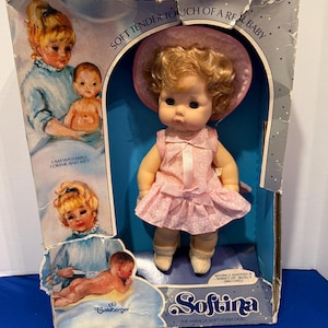 May include: A vintage Softina doll in its original box. The doll has blonde curly hair, a pink dress, and a matching hat. The box features illustrations of a baby and text that reads "Soft Tender Touch of a Real Baby."