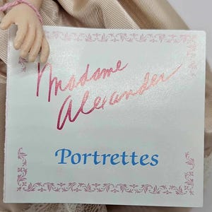 May include: A close-up of a tag with the words "Madame Alexander" in pink script above the word "Portrettes" in blue. The tag has a pink floral border and is held by a doll.