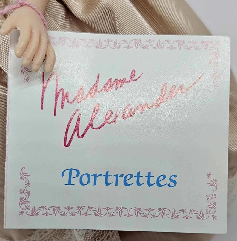 May include: A close-up of a tag with the words "Madame Alexander" in pink script above the word "Portrettes" in blue. The tag has a pink floral border and is held by a doll.