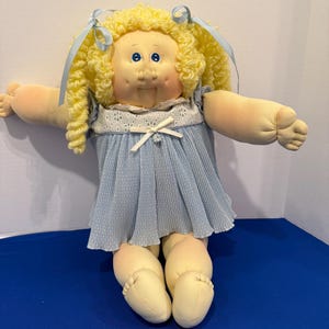 Può includere: Una bambola vintage con un corpo morbido e un viso rotondo. La bambola ha capelli biondi ricci legati con nastri blu, occhi blu e un vestito azzurro con finiture bianche. Le braccia e le gambe della bambola sono estese.