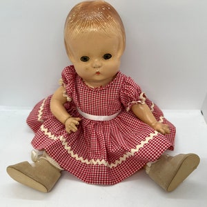 May include: A vintage doll with blonde hair, wearing a red and white gingham dress with white lace trim and brown shoes.