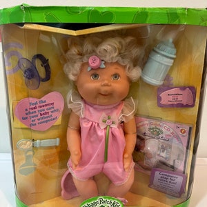 May include: Cabbage Patch Kids Cuddle 'n Care baby doll in a pink outfit with a blue bottle, a pacifier, and a CD-ROM. The box has a green background with a Cabbage Patch Kids logo.