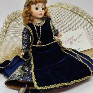 May include: A Madame Alexander doll wearing a dark blue velvet dress with gold trim and a matching headpiece. The doll has red hair, green eyes, and a gold necklace with a red pendant. The doll is wearing matching blue shoes.