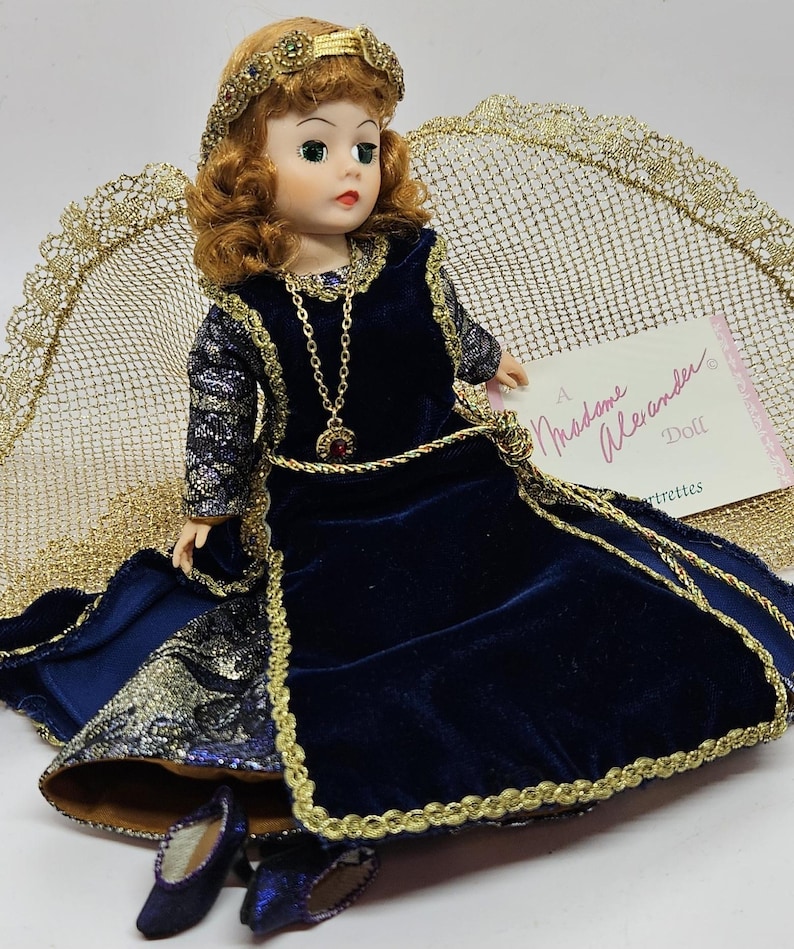 May include: A Madame Alexander doll wearing a dark blue velvet dress with gold trim and a matching headpiece. The doll has red hair, green eyes, and a gold necklace with a red pendant. The doll is wearing matching blue shoes.