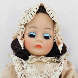 May include: A vintage doll with a painted face, blue eyes, and dark hair. The doll wears a cream-colored lace bonnet with pink ribbons and a matching lace collar. The doll's cheeks are blushed, and the lips are painted a soft orange.