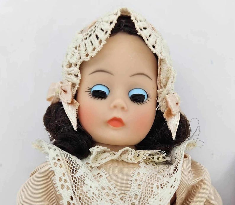 May include: A vintage doll with a painted face, blue eyes, and dark hair. The doll wears a cream-colored lace bonnet with pink ribbons and a matching lace collar. The doll's cheeks are blushed, and the lips are painted a soft orange.