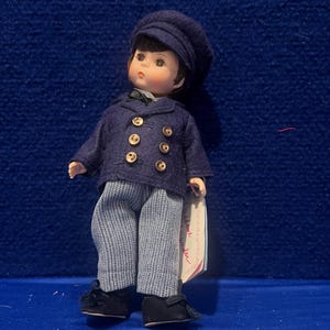 May include: A vintage doll of a young boy, dressed in a navy blue double-breasted jacket with gold buttons, striped pants, and a matching cap. The doll has black shoes and a small tag. The doll is standing on a blue surface.