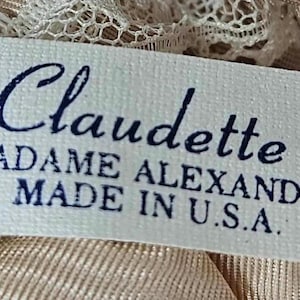May include: A rectangular label with the name "Claudette" in cursive, by Madame Alexander, made in the U.S.A. The label is white with dark blue text and is surrounded by beige fabric and lace.