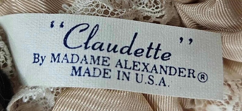 May include: A rectangular label with the name "Claudette" in cursive, by Madame Alexander, made in the U.S.A. The label is white with dark blue text and is surrounded by beige fabric and lace.