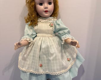 May include: A doll with wavy ginger hair and blue eyes wearing a light blue dress with a white checkered apron. The apron features small pink floral embroidery and lace trim. The doll has white tights and black shoes with a strap across the top.