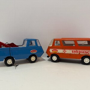 May include: Two vintage toy vehicles: a blue tow truck with a red tow arm and an orange emergency van. The emergency van has the word "EMERGENCY" and a white cross on the side. Both toys have black tires.