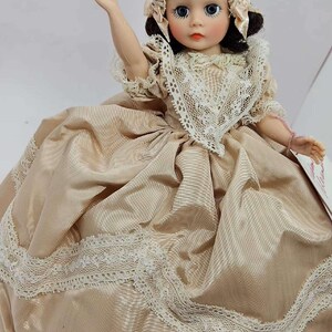 May include: A vintage doll wearing a beige satin dress with white lace trim. The doll has blue eyes, rosy cheeks, and a matching lace bonnet. The doll's arm is raised, and it appears to be seated. A classic collectible doll.