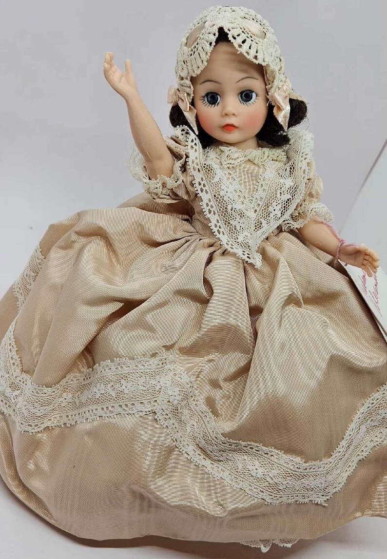 May include: A vintage doll wearing a beige satin dress with white lace trim. The doll has blue eyes, rosy cheeks, and a matching lace bonnet. The doll's arm is raised, and it appears to be seated. A classic collectible doll.