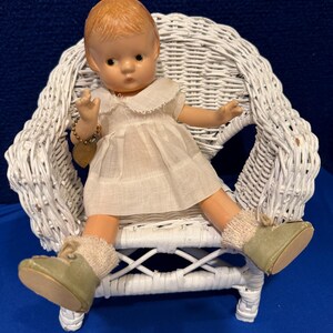 May include: A vintage doll with red hair, wearing a white dress, sitting in a white wicker chair. The doll has green shoes and beige socks. A gold-colored heart charm hangs from its wrist. The chair is on a blue surface.