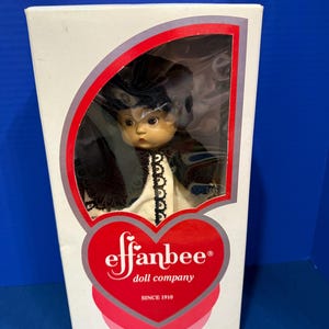Peut inclure: Une poupée Effanbee Doll Company dans une boîte blanche avec une fenêtre en plastique transparent. La poupée porte un chapeau sombre et une robe blanche avec une bordure en dentelle noire. La boîte présente un logo cœur rouge avec le texte Effanbee doll company SINCE 1910.