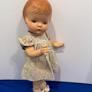 Könnte beinhalten: Vintage Puppe mit rotem Haar, grünen Augen und einem Kleid mit Blumenmuster. Die Puppe trägt weiße Schuhe mit Riemen. Das Kleid der Puppe hat eine blaue Bordüre. Die Puppe steht auf einer blauen Oberfläche.