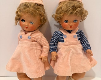 May include: Two vintage dolls wearing pink corduroy jumpers and matching hats. One doll has a striped shirt, the other has a blue and white striped shirt. Both dolls have blonde curly hair, rosy cheeks, and white shoes.