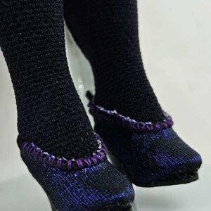 May include: Close-up of black doll stockings and shoes. The shoes are black with a purple and white trim. The shoes have a slight heel. The stockings are black.