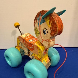 Könnte beinhalten: Vintage Ziehspielzeug Esel mit Patchwork-Design. Das Spielzeug hat einen gelben Körper, blaue Räder und eine rote Schnur. Der Kopf des Esels hat blaue Ohren und einen gelben Knopf.