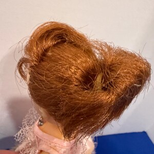 May include: A doll with auburn hair styled in a bun. The doll is wearing a pink dress with lace detailing. The doll's skin is a light tone, and the background is a mix of white and blue.
