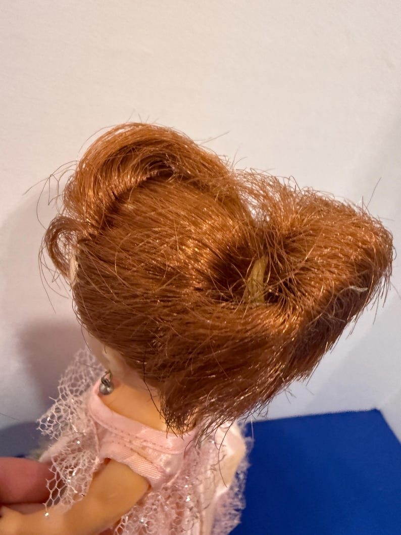 May include: A doll with auburn hair styled in a bun. The doll is wearing a pink dress with lace detailing. The doll's skin is a light tone, and the background is a mix of white and blue.