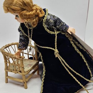 May include: A doll with auburn hair wearing a dark blue velvet gown with gold trim and accents. The doll is leaning towards a miniature wicker chair with a floral cushion. The doll's dress has gold rope detailing.