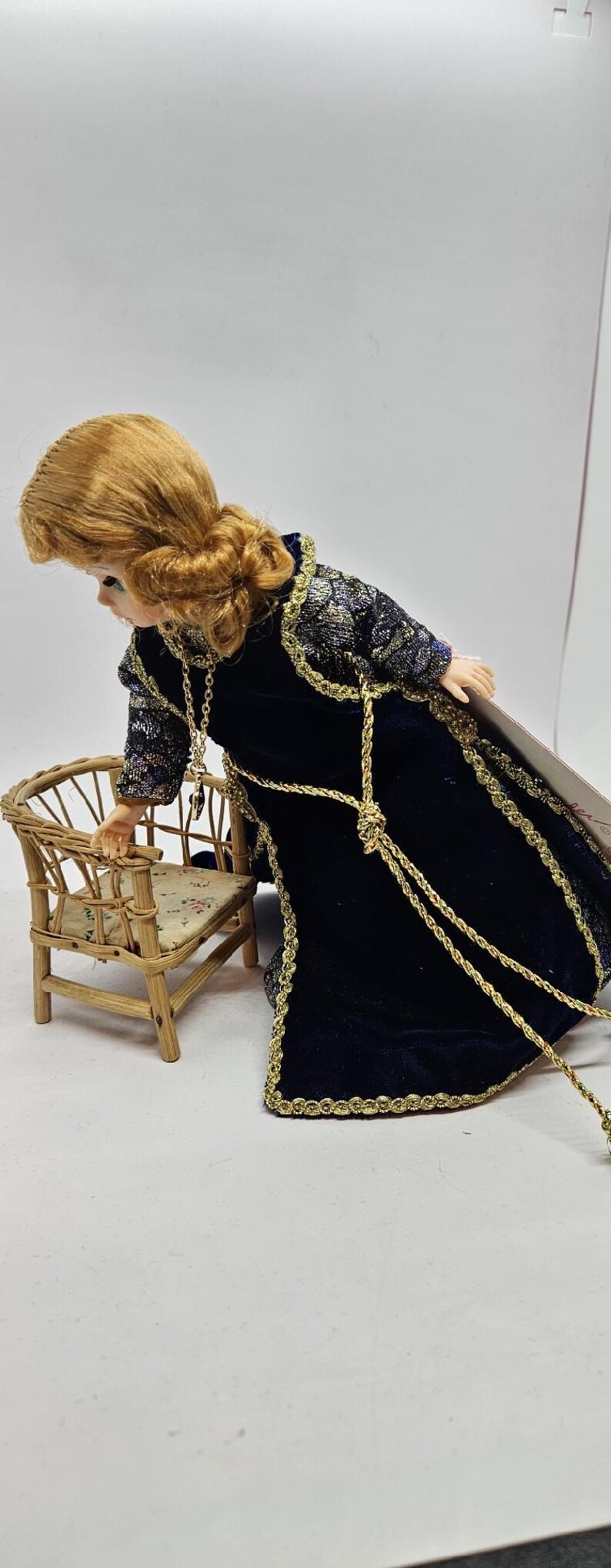 May include: A doll with auburn hair wearing a dark blue velvet gown with gold trim and accents. The doll is leaning towards a miniature wicker chair with a floral cushion. The doll's dress has gold rope detailing.