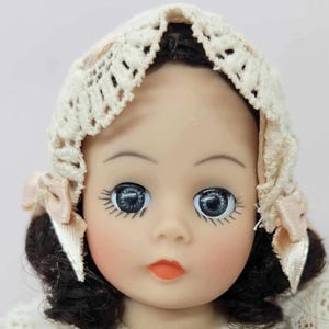 May include: A vintage doll with dark hair and large, blue eyes. The doll wears a cream-colored crocheted bonnet with pink ribbon accents. The doll has rosy cheeks and a small, orange-red mouth. The doll's face is smooth and porcelain-like.