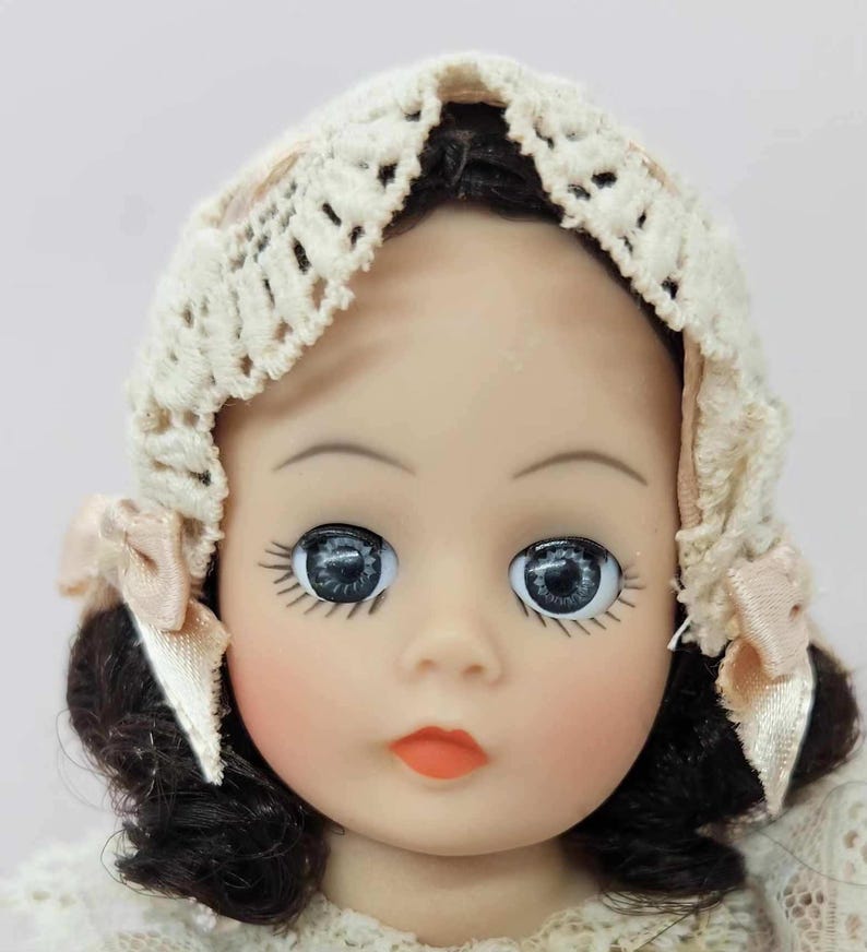 May include: A vintage doll with dark hair and large, blue eyes. The doll wears a cream-colored crocheted bonnet with pink ribbon accents. The doll has rosy cheeks and a small, orange-red mouth. The doll's face is smooth and porcelain-like.