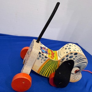 Può includere: Giocattolo vintage in legno a forma di cane da trascinare, con ruote arancioni e un manico nero. Il cane presenta un motivo a macchie, un orecchio nero e un cordino rosso. Il giocattolo è progettato con uno stile stravagante e fumettistico.