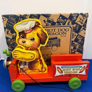 Può includere: Giocattolo vintage Fisher-Price "Hot Dog Wagon". Il carrello rosso presenta un cane dei cartoni animati chef e ruote verdi. La scatola dietro il carrello ha un motivo blu e marrone con le parole "Hot Dog Wagon".