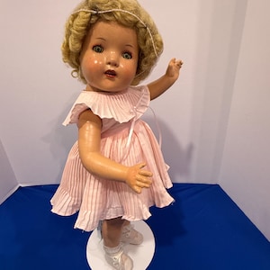 Peut inclure: Une poupée vintage avec des cheveux blonds bouclés et un nœud blanc. La poupée porte une robe plissée rose et des chaussures blanches. Elle est posée sur une base circulaire blanche sur fond bleu. La poupée a des joues roses et un bras levé.