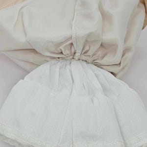 May include: A vintage doll dress, featuring a layered design. The dress has a white skirt with a delicate lace trim at the hem. The bodice is a soft cream color, and the lining is a light beige. The dress is gathered at the waist.