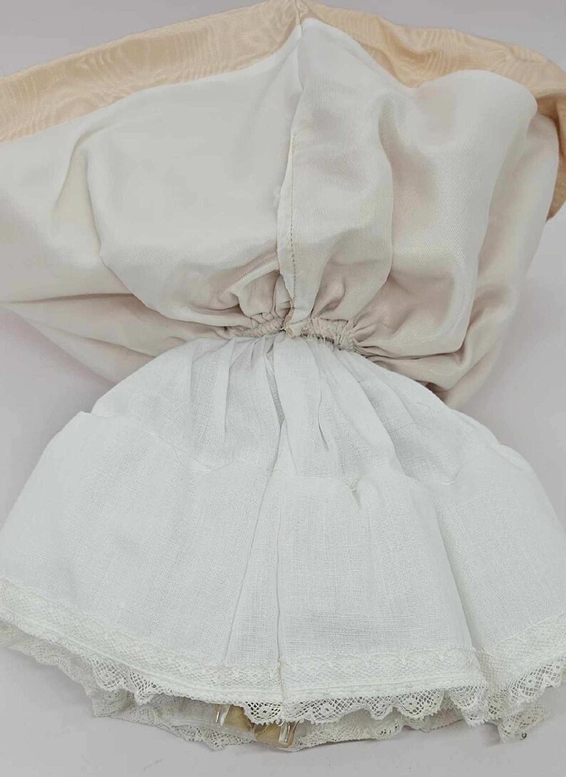 May include: A vintage doll dress, featuring a layered design. The dress has a white skirt with a delicate lace trim at the hem. The bodice is a soft cream color, and the lining is a light beige. The dress is gathered at the waist.