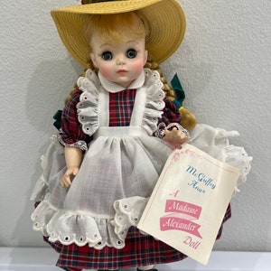 May include: A Madame Alexander doll wearing a red and black plaid dress, a white apron, and a straw hat with flowers. The doll is holding a tag that reads "McGuffey Ana® A Madame Alexander Doll".