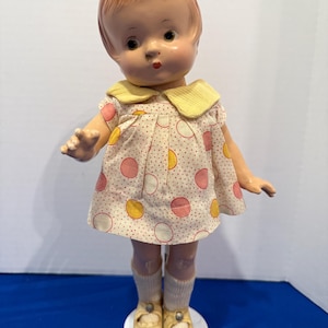 May include: Vintage doll with red hair, wearing a white dress with pink and yellow polka dots and a yellow collar. The doll has a painted face, socks, and yellow shoes. The doll is standing on a white base.