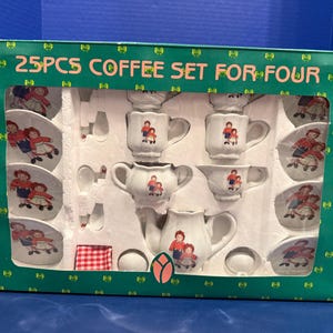 Peut inclure: Un ensemble à café de 25 pièces pour quatre personnes dans une boîte verte avec fenêtre. Le service en céramique blanche comprend une théière, un pot à lait, un sucrier, des tasses, des soucoupes et des cuillères, décorés de deux poupées. Texte : 25PCS COFFEE SET FOR FOUR.